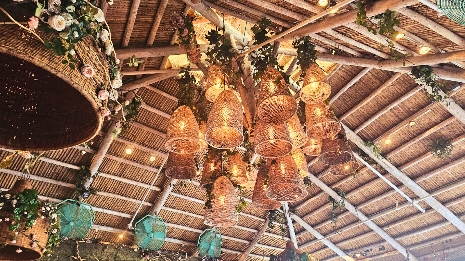 Interior view of a thatched roof Tiki Hut with exposed wooden beams and a cluster of woven hanging light fixtures and string lights.