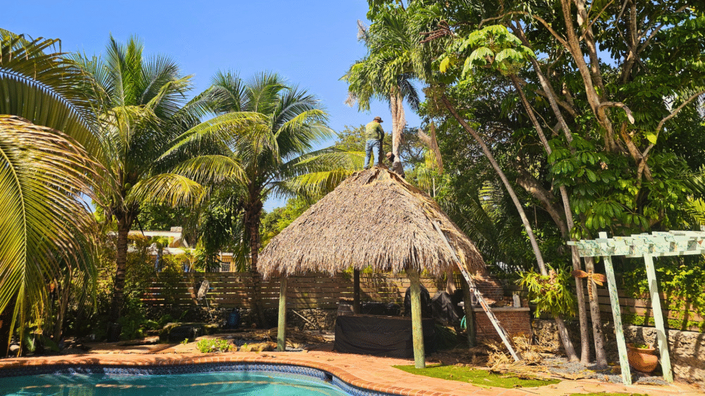 Island Tiki Hut construction by the poolside with a worker installing the thatched roof in a tropical backyard.
